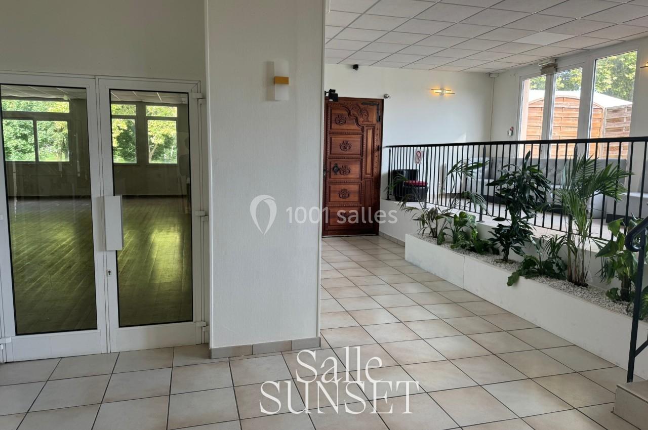 Entrée d'une salle avec sol carrelé, plantes en intérieur, portes vitrées à gauche et grande porte en bois au fond.