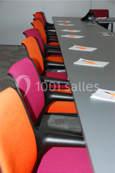 Chaises colorées alignées le long d'une table grise avec des carnets et stylos orange posés devant chaque siège.