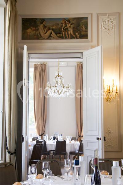 Salle de restaurant élégante avec lustre, moulures, rideaux beiges et peinture décorative au-dessus des portes.