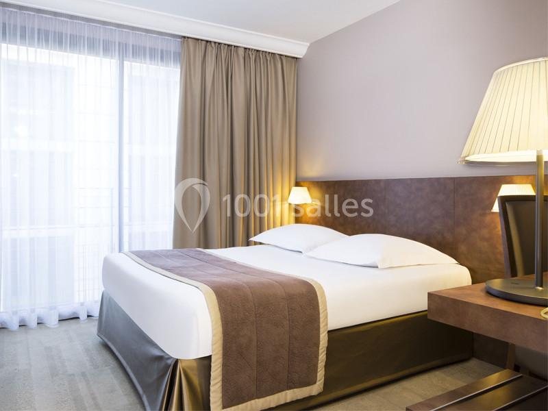 Chambre d'hôtel lumineuse avec un lit double, des draps blancs, des rideaux beiges et une lampe de chevet allumée.