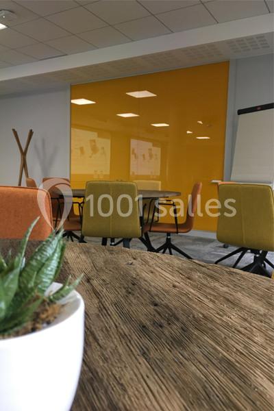 Salle de réunion moderne avec table en bois, chaises colorées, mur jaune, tableau de conférence et plante au premier plan.