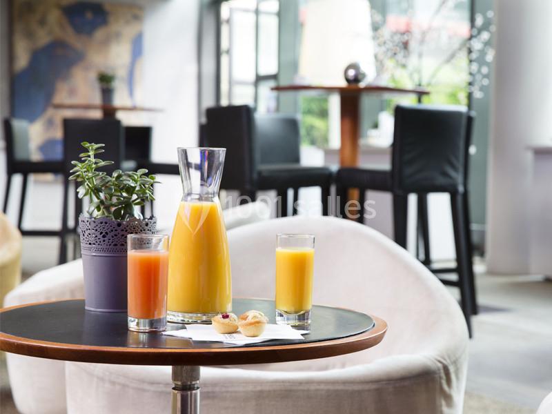 Carafe de jus d'orange, verres de jus et petites pâtisseries sur une table ronde dans un espace lounge lumineux.