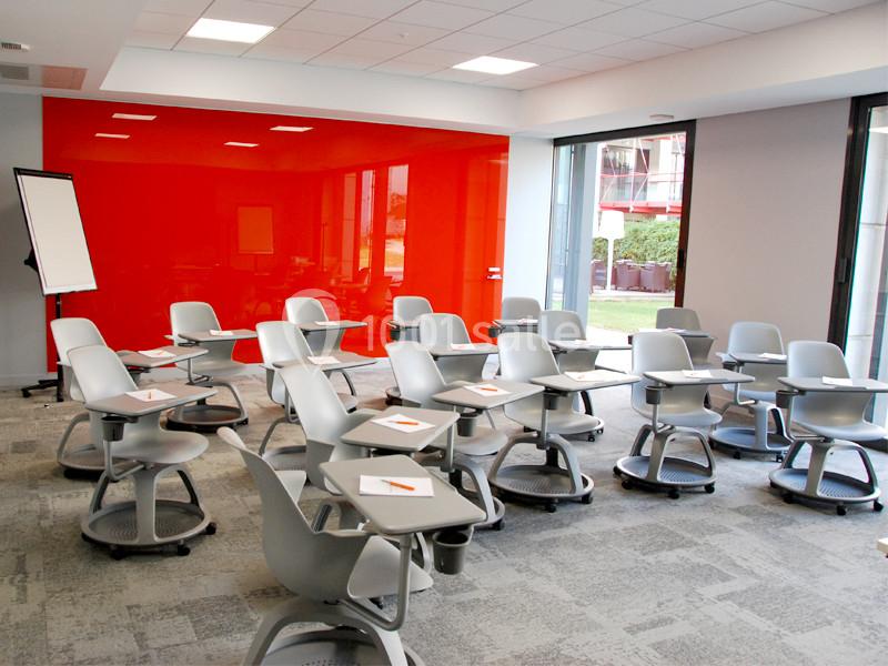 Salle de classe moderne avec des chaises équipées de tablettes, un mur rouge vif et un tableau de conférence.
