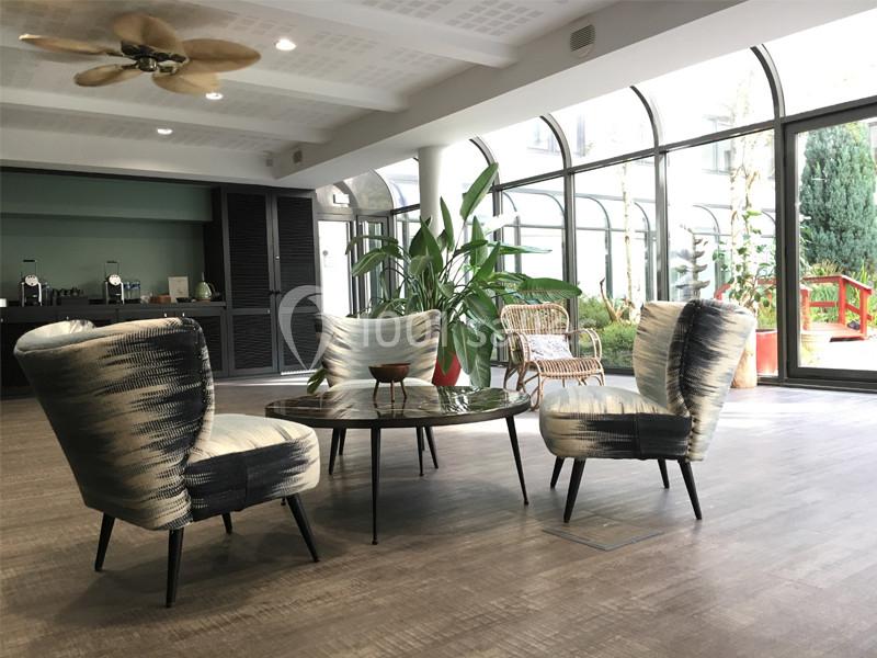 Espace salon lumineux avec fauteuils modernes, table basse en verre, plantes et grandes baies vitrées donnant sur un jardin.