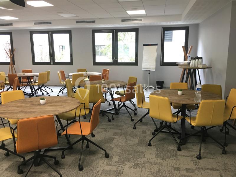 Salle de réunion lumineuse avec tables rondes en bois, chaises colorées et tableau de présentation.