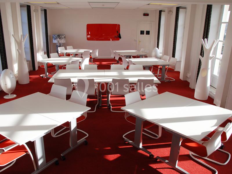 Salle lumineuse avec tables blanches, chaises modernes et sol rouge, aménagée pour le travail ou les réunions.