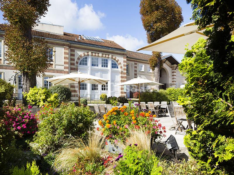 Jardin fleuri avec terrasse aménagée, tables et parasols devant une bâtisse en briques et pierres sous un ciel bleu.