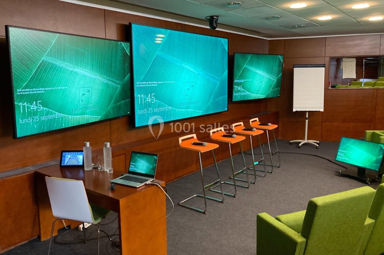 Salle de conférence équipée d'écrans, de tabourets, d'un tableau blanc, et d'un bureau avec un ordinateur portable.