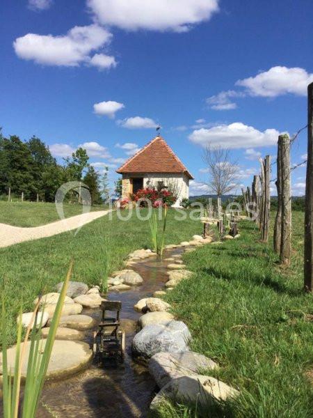 Petit ruisseau bordé de pierres menant à une petite maison en pierre avec un toit rouge, sous un ciel bleu.