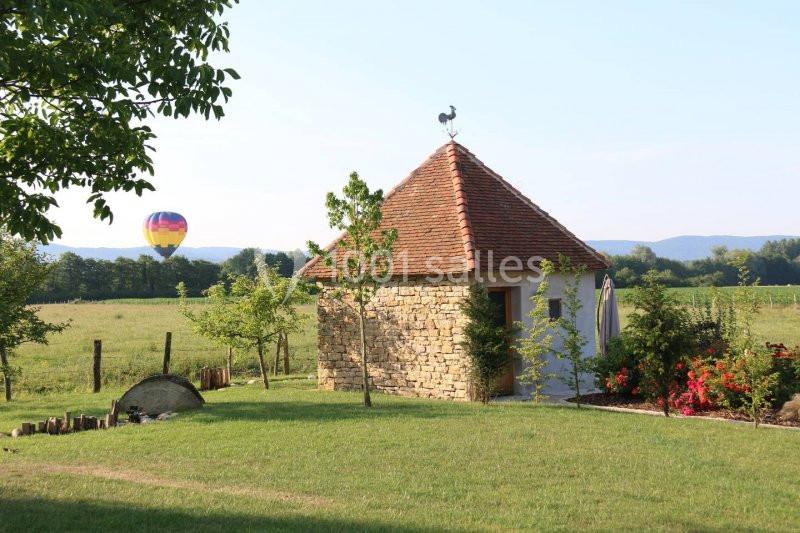 Petite maison en pierre avec toit en tuiles, entourée de verdure, une montgolfière visible dans le ciel en arrière-plan.