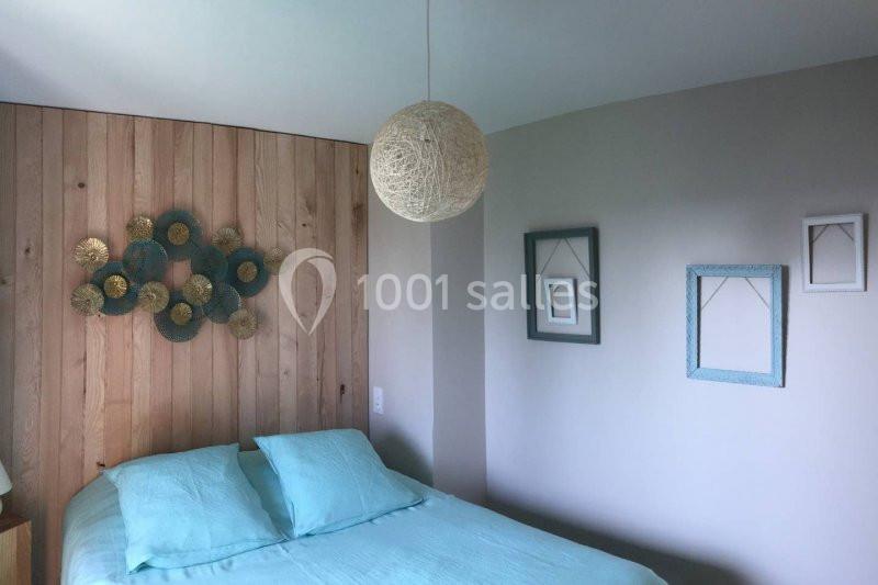 Lit double avec draps bleus, mur en bois décoré d'un ornement circulaire et deux cadres bleus sur un mur blanc.