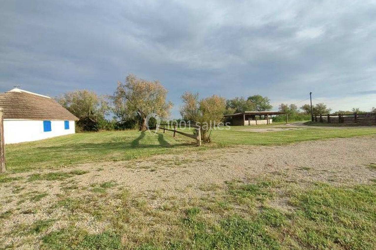 Terrain herbeux avec une maison blanche aux volets bleus, une clôture en bois et un abri sous un ciel nuageux.