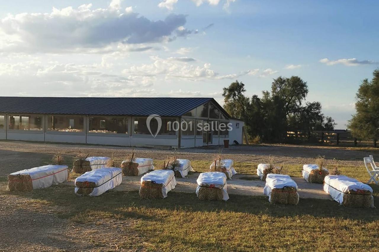 Bottes de paille recouvertes de tissu blanc disposées en rangées devant un bâtiment avec toit en tôle, sous un ciel dégagé.