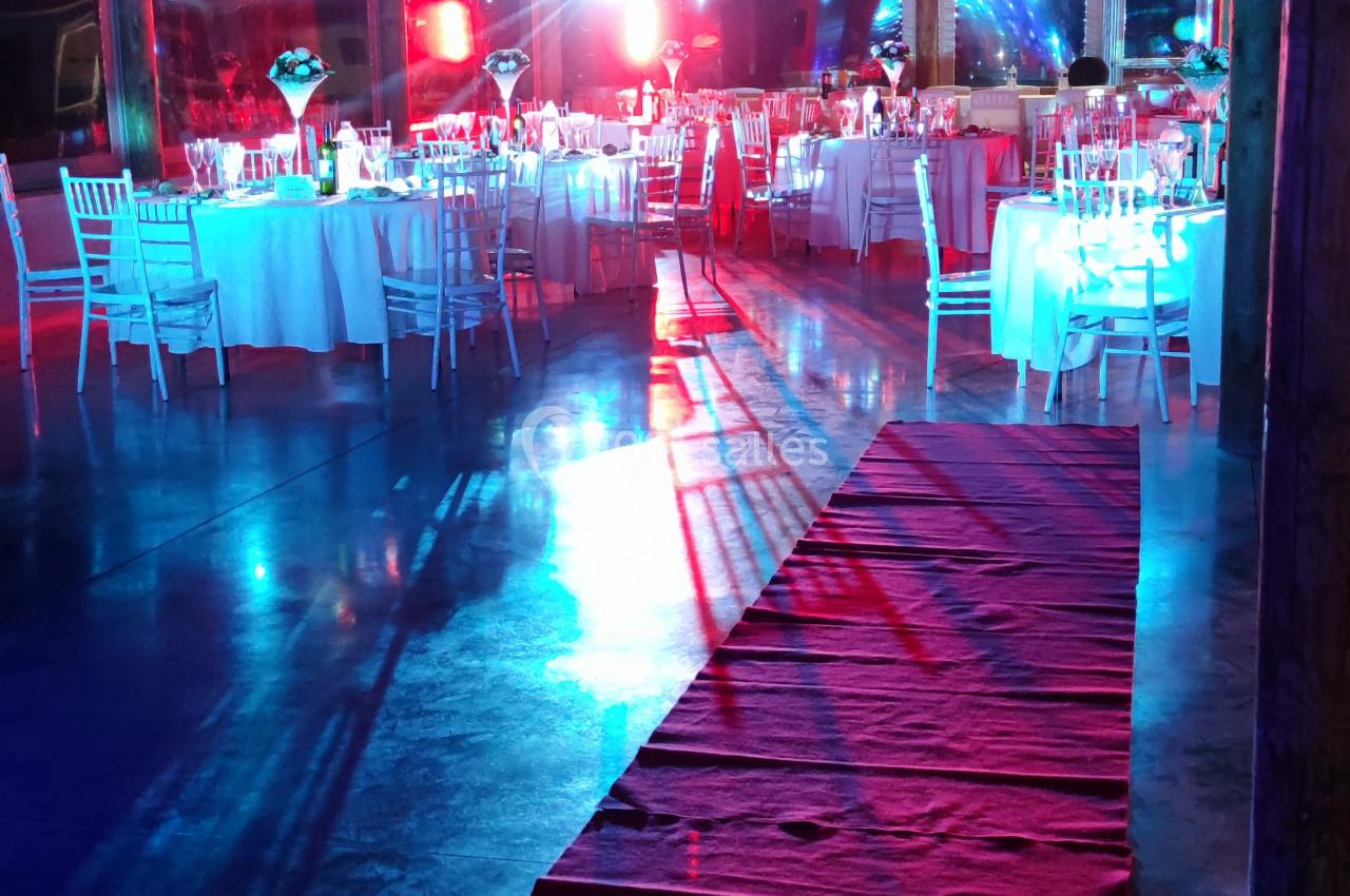 Salle de réception avec tables dressées, éclairages colorés et tapis rouge menant à la scène.
