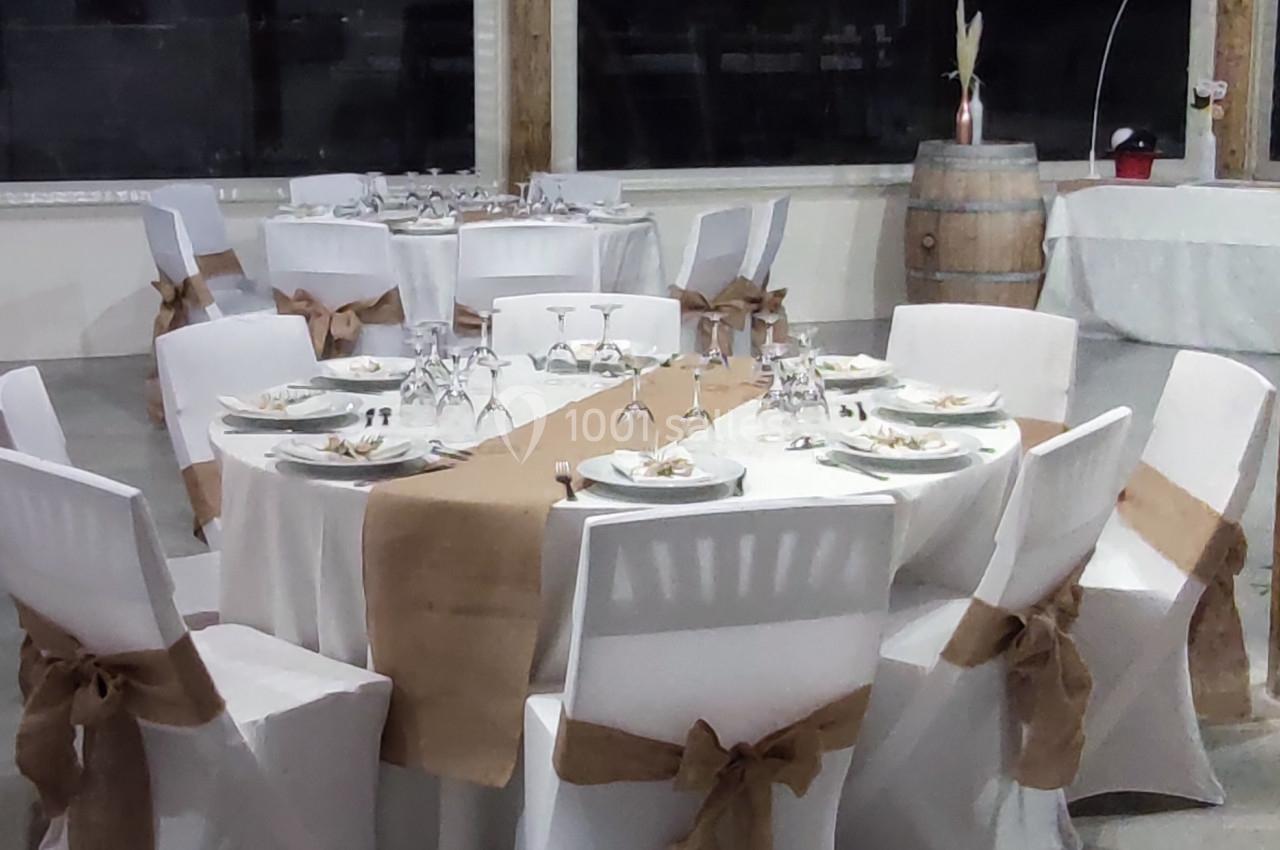 Salle décorée pour un événement avec tables rondes dressées, nappes blanches et chaises ornées de nœuds en toile de jute.