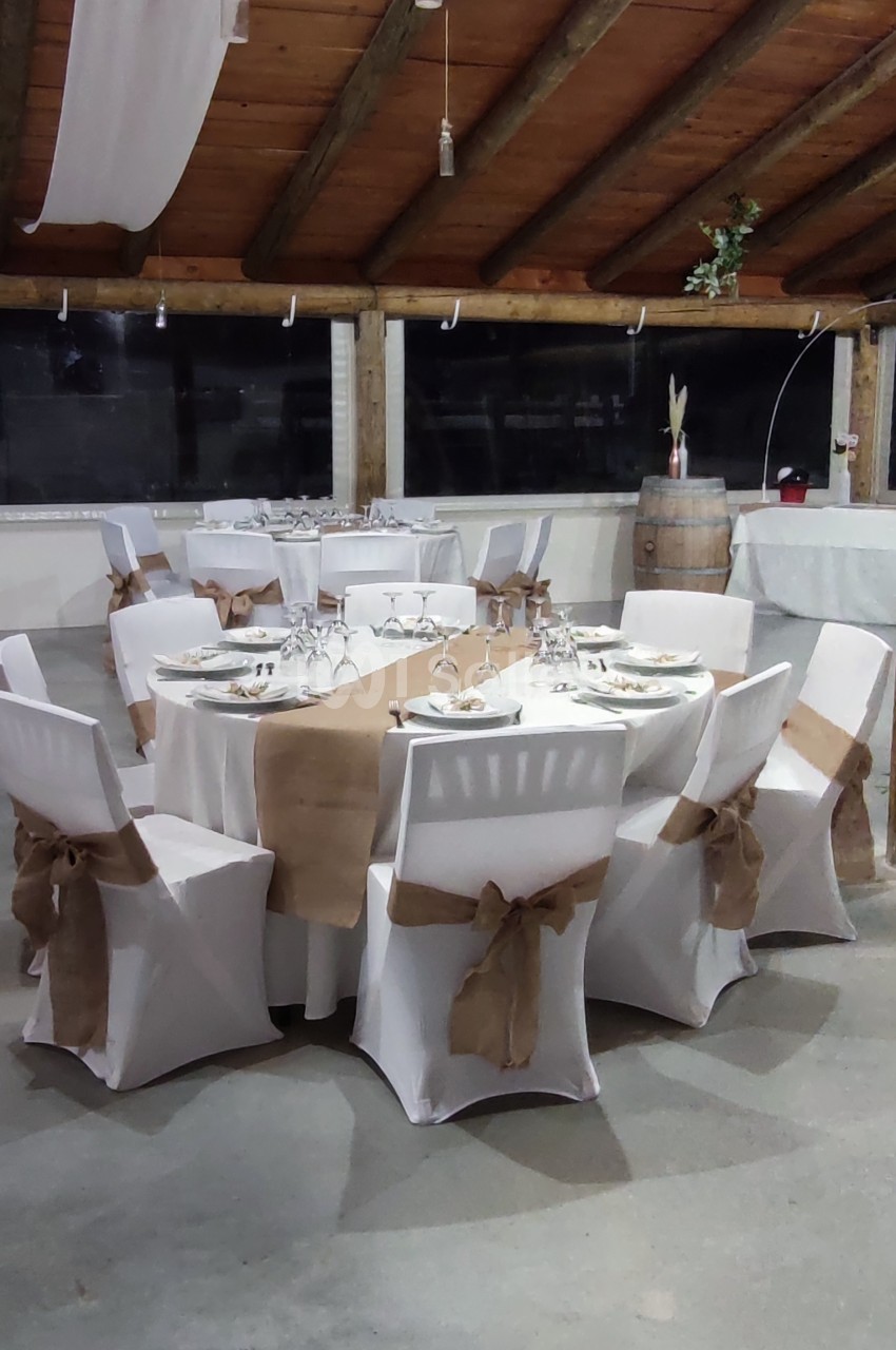 Salle décorée pour un événement avec tables rondes dressées, nappes blanches et chaises ornées de nœuds en toile de jute.