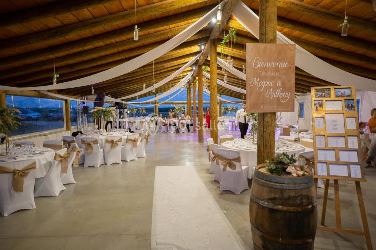 Salle de réception décorée pour un mariage, avec tables dressées, guirlandes suspendues et panneau de bienvenue en bois.