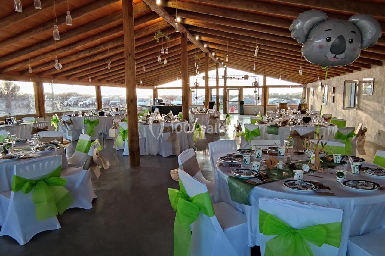 Salle de réception décorée pour un événement, avec des tables dressées et des chaises ornées de nœuds verts.