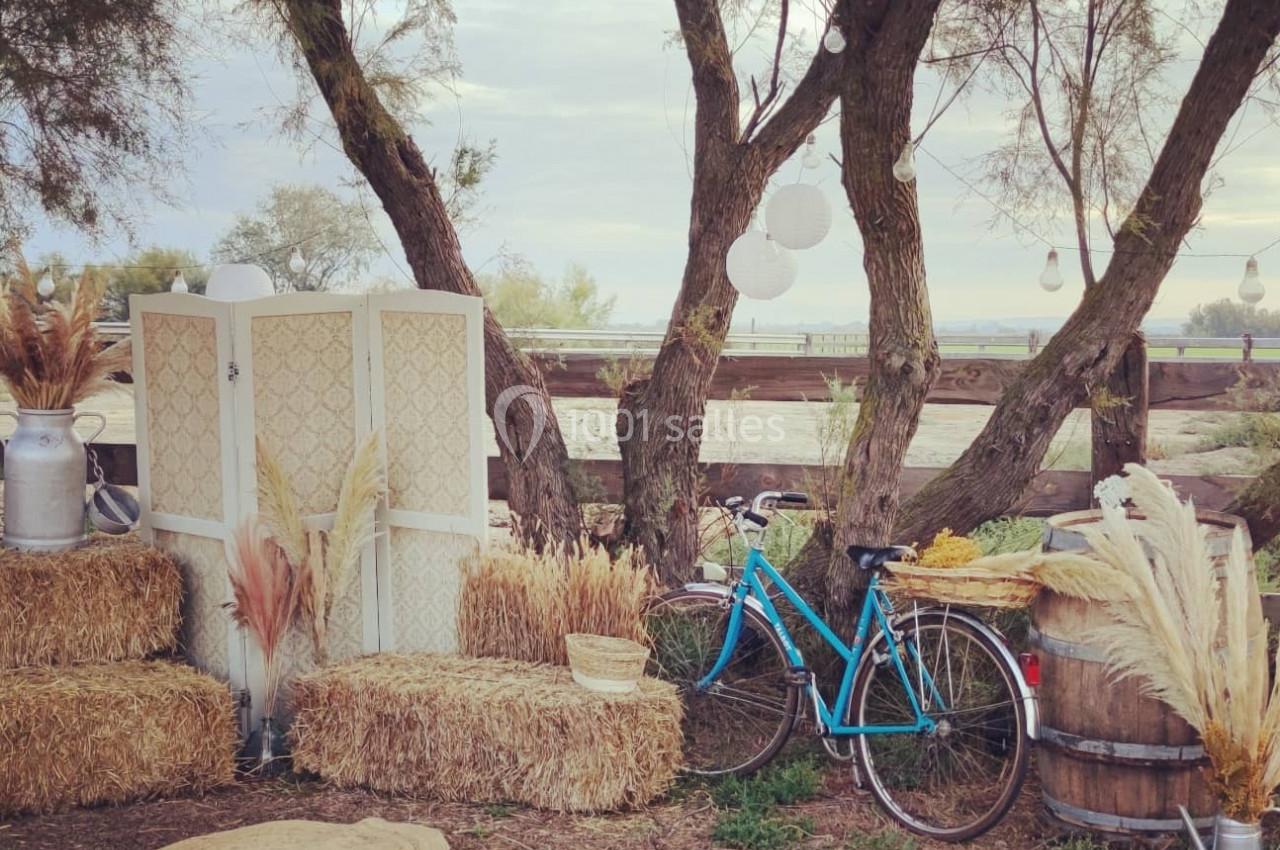 Décor champêtre avec bottes de paille, vélo bleu, plantes séchées, tonneaux en bois et arbres en arrière-plan.