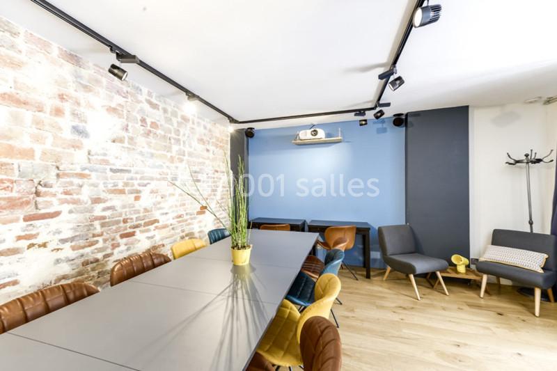 Salle lumineuse avec mur en briques, table longue entourée de chaises colorées, fauteuils et projecteur au mur.