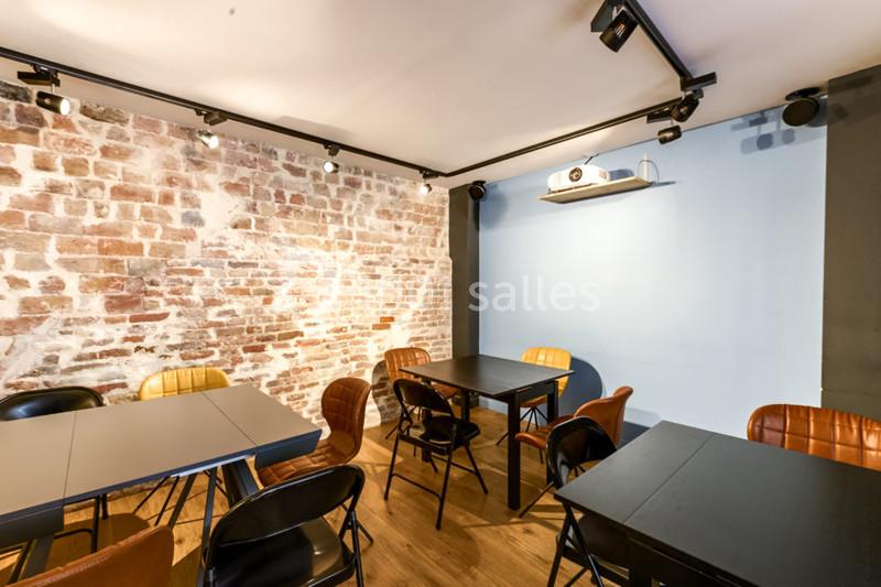 Salle lumineuse avec murs en briques et peinture bleue, tables noires, chaises variées et projecteur au plafond.