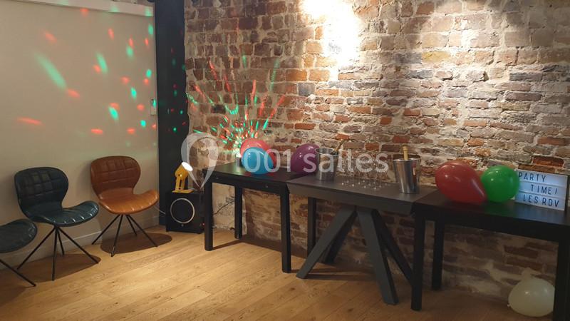 Salle décorée pour une fête avec des ballons colorés, des tables garnies de verres et un mur en briques apparentes.