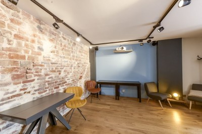 Salle de réunion moderne avec table noire, chaises, écran de projection, paperboard et cuisine ouverte en arrière-plan.