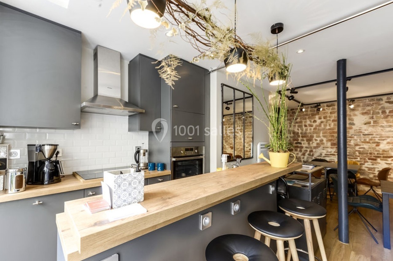 Cuisine moderne avec comptoir en bois, tabourets noirs, murs en briques apparentes et éclairage suspendu.