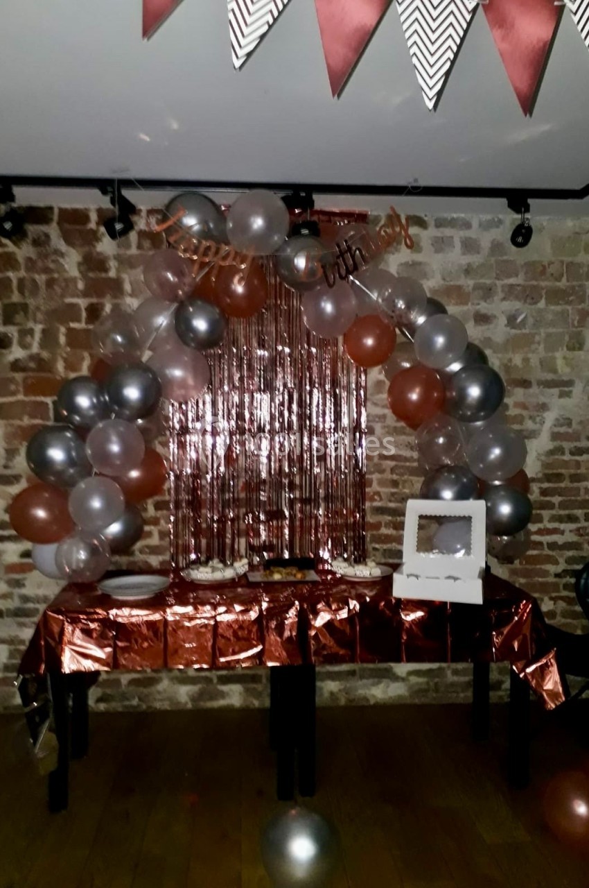 Décoration d'anniversaire avec ballons argentés et cuivrés, guirlande et table dressée devant un mur de briques.
