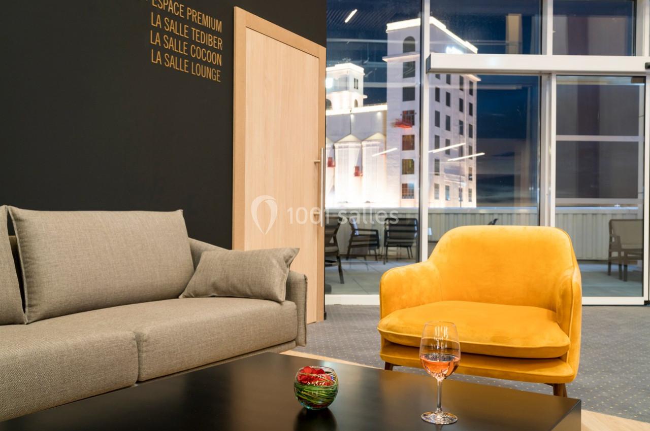 Salon moderne avec canapé gris, fauteuil jaune, table basse noire et vue sur une terrasse éclairée en arrière-plan.