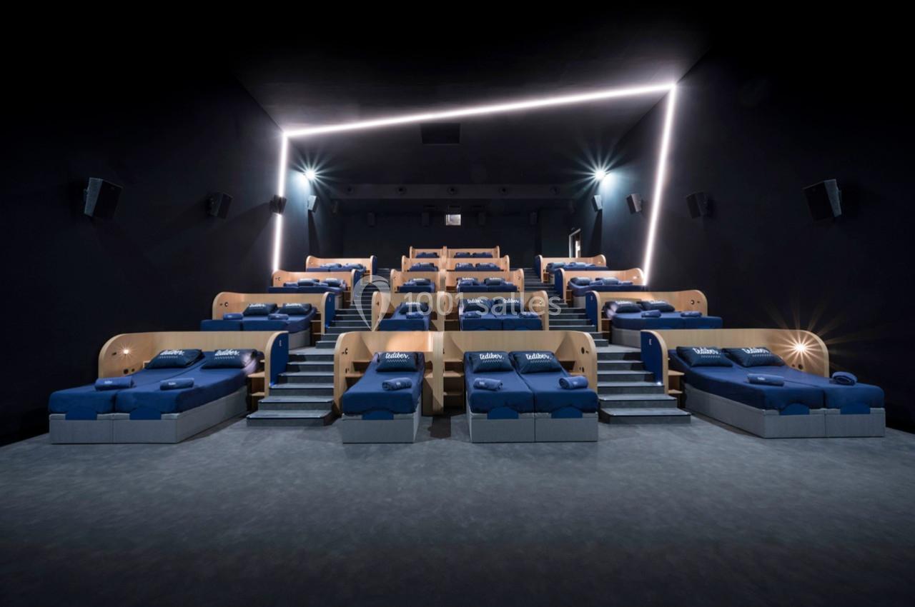Salle de cinéma avec des lits bleus alignés en gradins, éclairage tamisé et projecteurs muraux.
