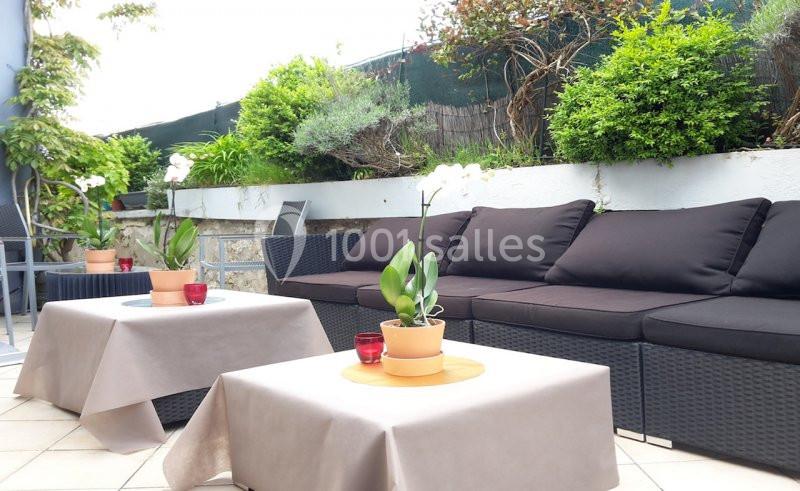 Salon de jardin avec canapé en rotin noir, tables basses couvertes de nappes beige et plantes en pots sur une terrasse.