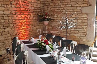 Cour d'une maison en pierre avec terrasse fleurie, arbres, guirlandes lumineuses et sol en gravier.