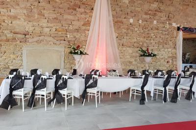 Cour d'une maison en pierre avec terrasse fleurie, arbres, guirlandes lumineuses et sol en gravier.