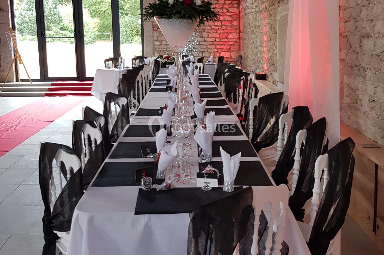 Salle de réception avec une longue table dressée, nappes blanches, chaises décorées de nœuds noirs et centre de table floral.