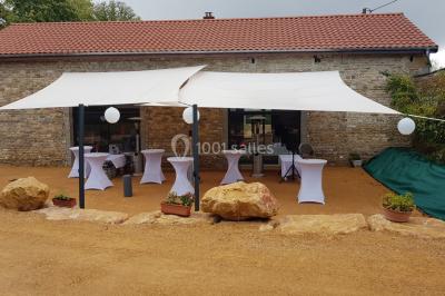 Cour d'une maison en pierre avec terrasse fleurie, arbres, guirlandes lumineuses et sol en gravier.