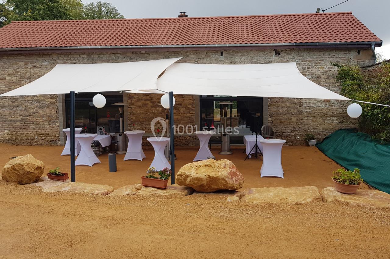Terrasse aménagée avec des voiles d'ombrage, tables hautes habillées de housses blanches et décorations extérieures.