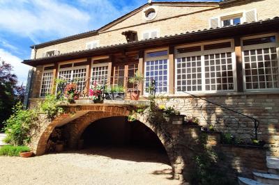 Cour d'une maison en pierre avec terrasse fleurie, arbres, guirlandes lumineuses et sol en gravier.
