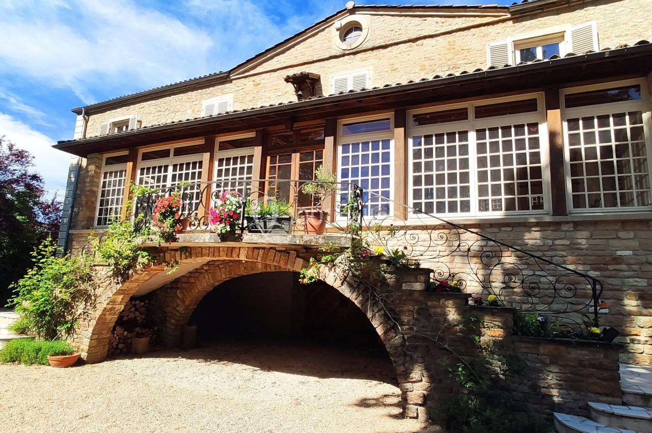 Façade d'une maison en pierre avec une terrasse surélevée, des fenêtres à carreaux et un escalier en fer forgé.