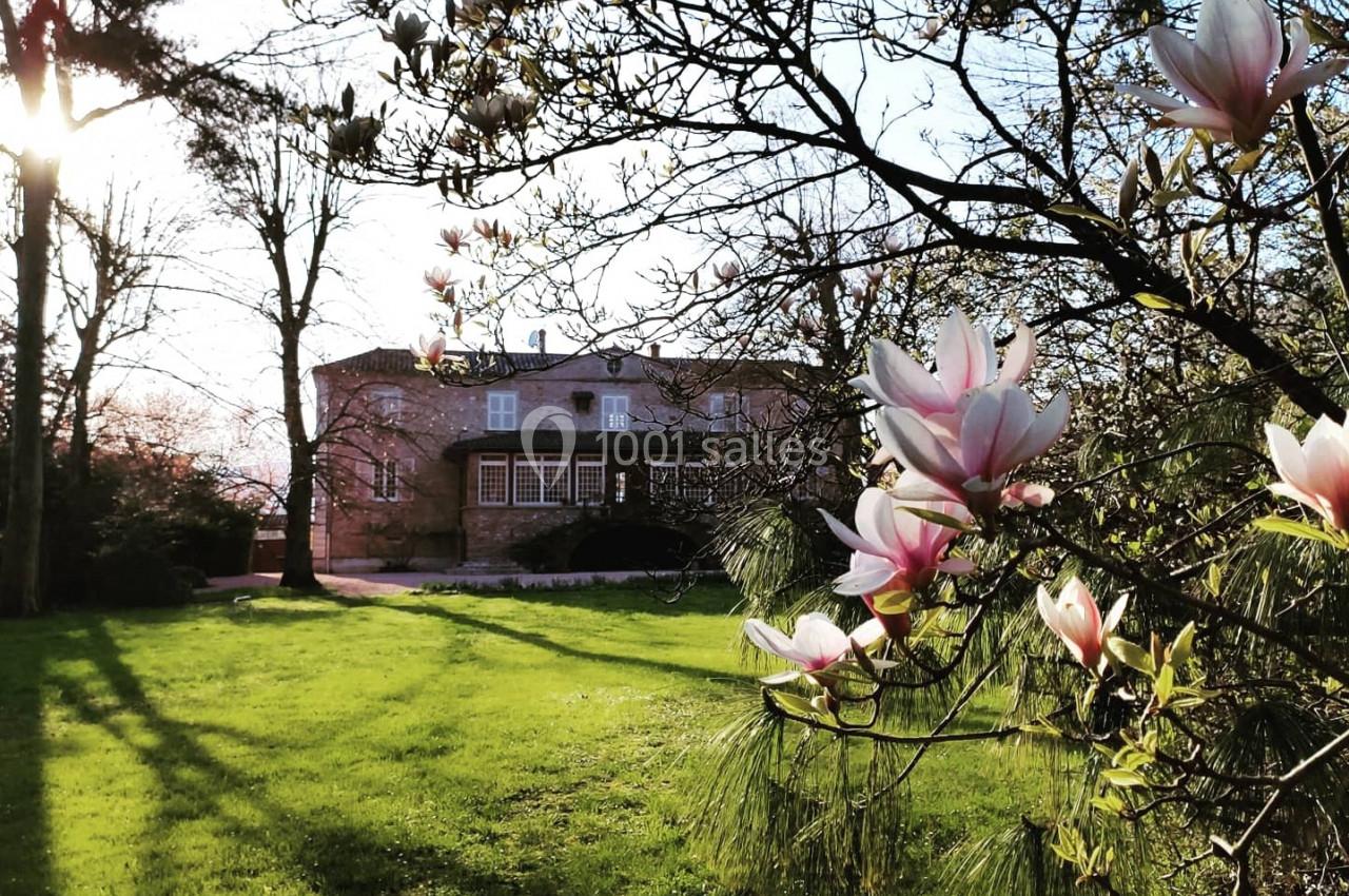 Maison en pierre entourée d'un jardin verdoyant, avec des magnolias en fleurs au premier plan et un soleil couchant.