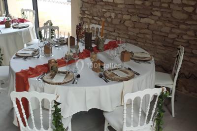 Cour d'une maison en pierre avec terrasse fleurie, arbres, guirlandes lumineuses et sol en gravier.