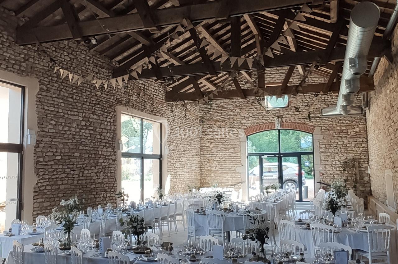 Salle de réception en pierre avec tables dressées, chaises blanches et décoration florale sous un plafond en bois.