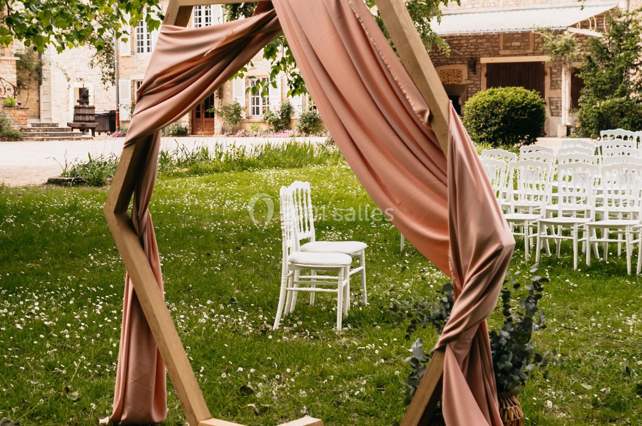Arche hexagonale en bois décorée de drapés roses, installée sur une pelouse devant des chaises blanches et un bâtiment en…