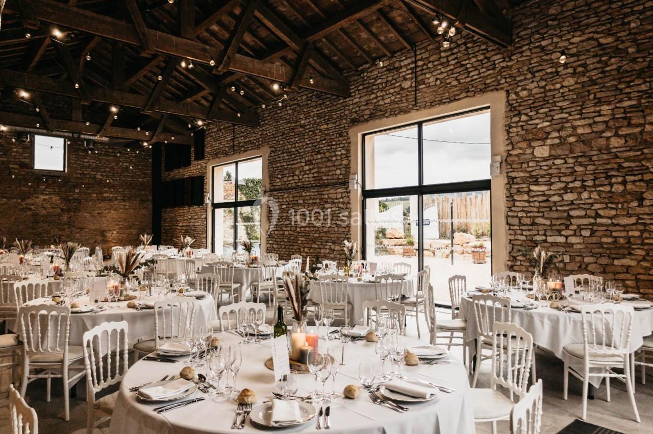 Salle de réception en pierre avec tables rondes dressées, chaises blanches et lumière naturelle entrant par de grandes baies…