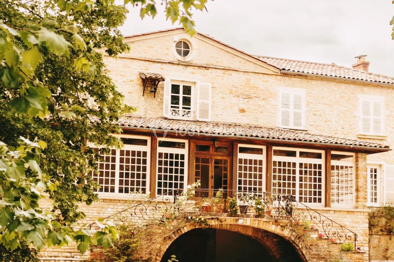 Façade d'une maison en pierre avec de grandes fenêtres, un porche voûté et des arbres au premier plan.