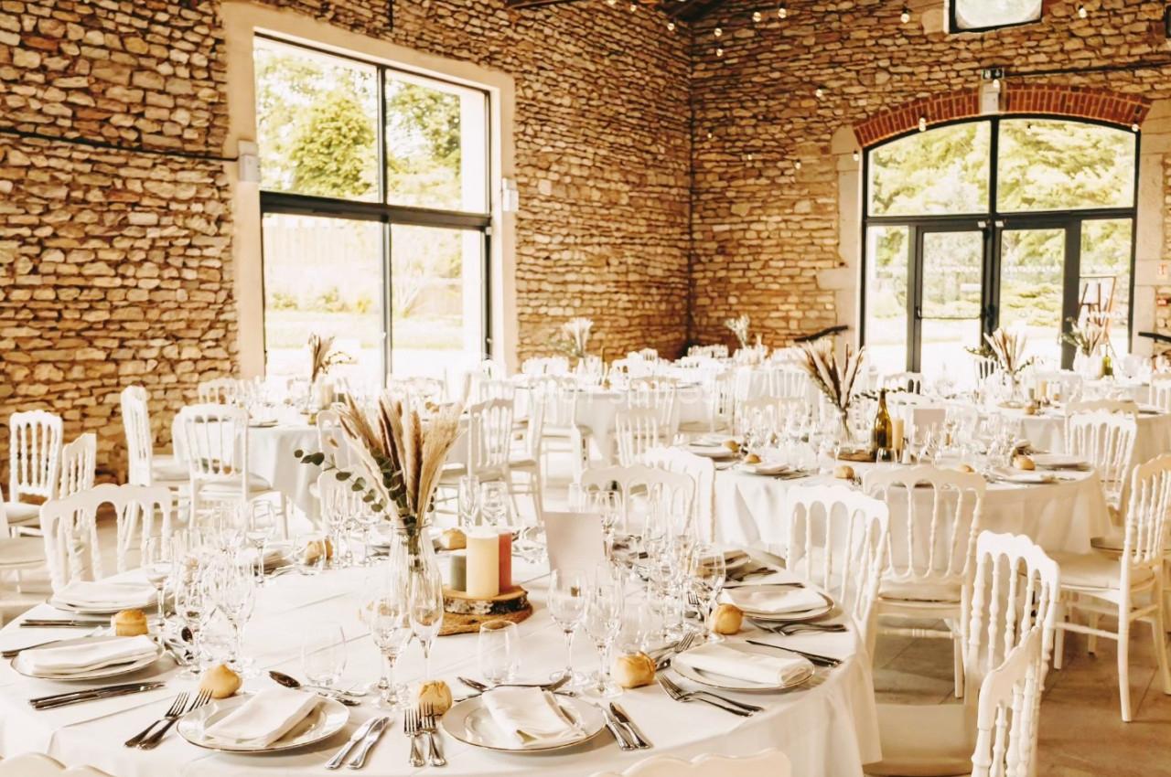 Salle de réception en pierres apparentes avec tables rondes dressées, décorées de bouquets et chaises blanches.