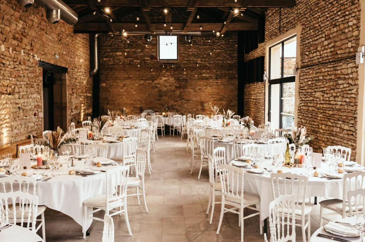 Salle en pierre avec plafond en bois, tables rondes dressées avec nappes blanches et chaises blanches, éclairage chaleureux.