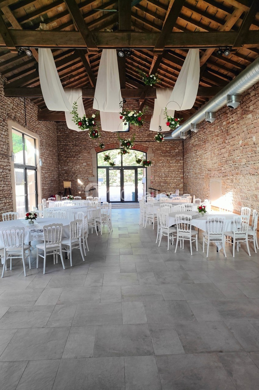 Salle de réception en pierre avec tables dressées, chaises blanches et décorations suspendues sous un plafond en bois.