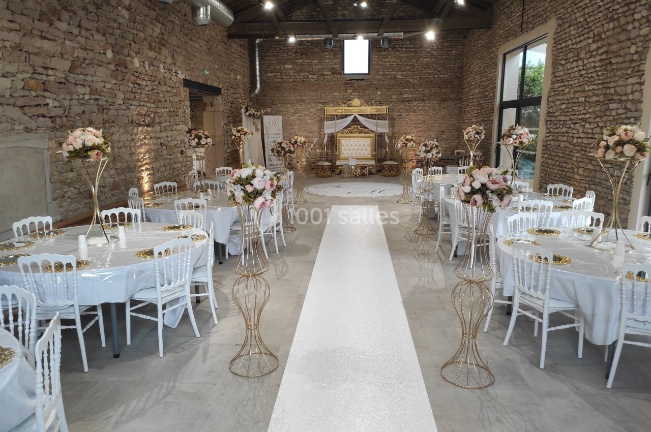 Salle de réception décorée avec des tables rondes, des chaises blanches et une allée centrale menant à une arche fleurie.