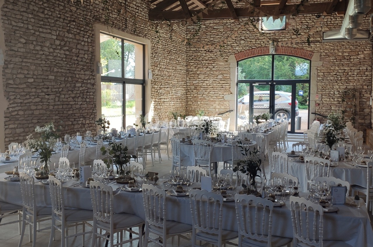 Salle de réception en pierre avec de longues tables décorées pour un événement, éclairée par de grandes baies vitrées.